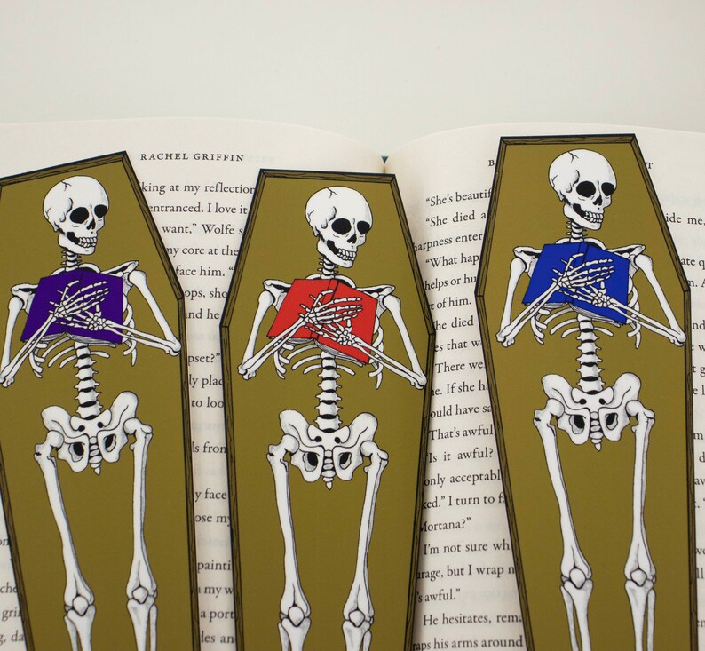 May include: Three skeleton bookmarks with different coloured books in their hands. The bookmarks are shaped like coffins and have a gold background. The text on the bookmarks reads "She's beautiful", "She died a", "harpness enter", "What hap", "helps or hu", "t of him", "he died", "es that w", "There we", "e. If she h", "ould have sa", "That's awful", "Is it awful?", "only acceptabl", "ked." I turn to f", "Mortana?", "I'm not sure wh", "arage, but I wrap n", "sawful.", "He hesitates, rem", "raps his arms around", "ide me", "ate o", "nd", "W", "m", "d", "av".