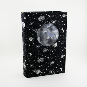 Puede incluir: Un diario de tapa dura con fondo negro cubierto de estrellas, galaxias y planetas blancos. Un gran gráfico detallado de la luna está centrado en la portada. El diseño del diario evoca un tema celestial, perfecto para escribir o dibujar.