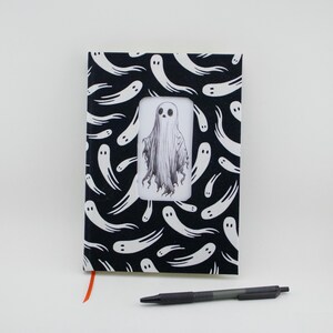 Puede incluir: Un cuaderno negro y blanco con un patrón de fantasmas. La portada presenta un solo fantasma más grande en el centro rodeado de fantasmas más pequeños. El cuaderno tiene un marcador de cinta roja.