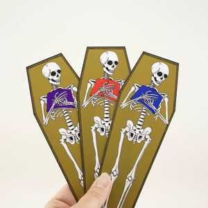 May include: Three gold-coloured coffins with skeletons holding books. The skeletons are wearing purple, red, and blue books. The coffins are standing upright.