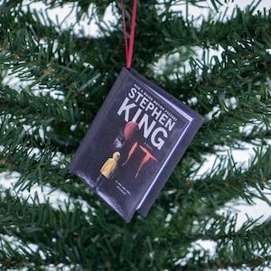 Peut inclure: Ornement de Noël en forme de livre miniature. La couverture affiche le titre "IT" de Stephen King, avec un ballon rouge et une figurine vêtue de jaune. L'ornement est suspendu par un ruban rouge, sur fond d'aiguilles de pin vertes.