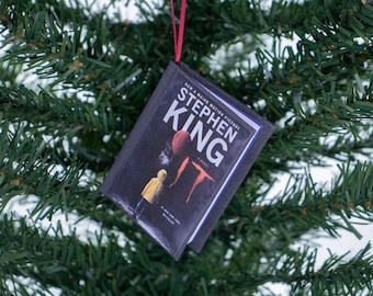 May include: A Christmas tree ornament shaped like a miniature book. The book cover features the title "IT" by Stephen King, with a red balloon and a yellow-clad figure. The ornament hangs from a red ribbon against a backdrop of green pine needles.