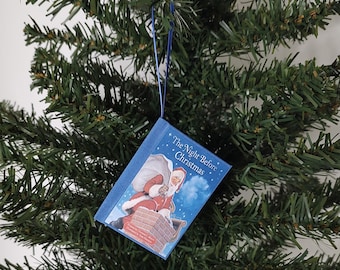 Pode incluir: Enfeite de árvore de Natal em forma de livro em miniatura, com a capa de "The Night Before Christmas". A ilustração mostra o Pai Natal num telhado. O enfeite é azul com texto branco e um cordão azul para pendurar.