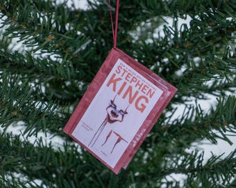 May include: A miniature book ornament featuring the cover of Stephen King's "IT" novel. The ornament has a red cover with the book title and a graphic of a clown's face. The ornament is hanging from a red string against a backdrop of green Christmas tree needles.