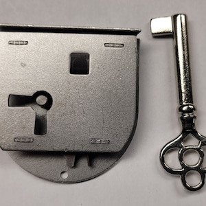 May include: A silver-colored antique lock and key set. The lock has a rectangular shape with a keyhole and a square opening. The ornate key is next to the lock. The lock and key are likely for a cabinet or small box.