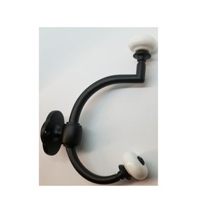 May include: Black metal coat hook with two white ceramic knobs. The hook is curved and has a rounded base.