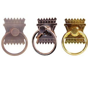 May include: Three decorative cabinet door pulls with a ring handle. The pulls are in three finishes: antique brass, black, and polished brass.