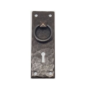 May include: A rustic, rectangular, metal door pull with a ring handle and a keyhole. The pull has a hammered finish and is a dark brown color.