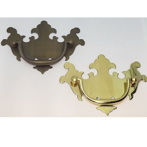 May include: Two ornate cabinet pulls, one in a brass finish and one in an antique brass finish. Both pulls have a decorative, scroll-like design.