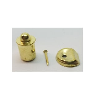 May include: A set of three gold-toned metal doorstop hardware pieces. The set includes a round doorstop with a hole in the center, a small nail, and a cylindrical doorstop with a rounded top.