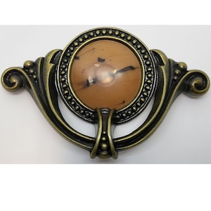 May include: A decorative cabinet knob with a brown and black marbled design. The knob is surrounded by a gold metal frame with a beaded edge.