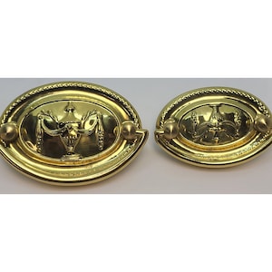 May include: Two gold-toned oval cabinet pulls with a decorative urn design. The pulls have a raised edge and a smooth, polished finish.