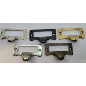 May include: Five drawer pulls in different finishes: nickel, black, brass, satin nickel, and antique brass. Each pull has a rectangular plate with a slot for a label and a rounded knob.