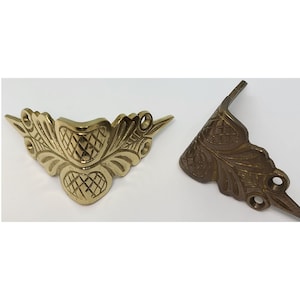 May include: Two ornate brass corner brackets with a leaf and acorn design. The bracket on the left is polished brass, while the bracket on the right is a more antique brass finish.