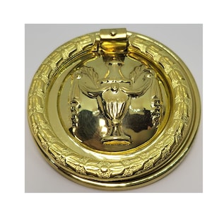 May include: Gold-toned metal door knob with a decorative urn design and a ring pull. The knob has a detailed, ornate border.