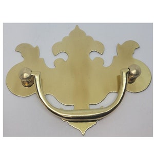 May include: A gold-toned metal drawer pull with a decorative, ornate design. The pull has a curved handle and two round knobs.