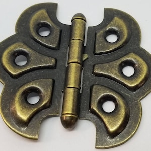 May include: A decorative butterfly-shaped hinge with a brass finish. The hinge has a detailed design with multiple rounded cutouts and a textured surface.