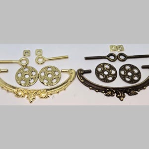 May include: Two sets of ornate metal drawer pulls, one in gold and one in black. Each set includes two decorative round pieces, two small square pieces, and a long, thin rod with a loop at each end.