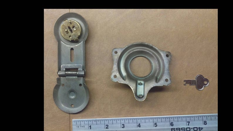 May include: A set of three antique metal lock parts, including a hinged lock with a brass keyhole, a circular lock plate with a keyhole, and a small key. The lock parts are on a brown surface with a ruler for scale.