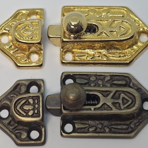 May include: Two decorative latches in gold and bronze tones. Each latch has an ornate design with a central locking mechanism and a matching catch. The latches are likely for a small box or cabinet, and are shown against a white background.