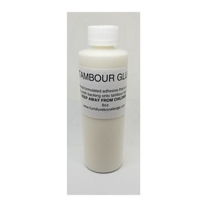 May include: A white plastic bottle of Tambour Glue, a special adhesive for tambour doors. The bottle is labeled with the text "Tambour Glue", "Special formulated adhesive that is ideal for applying cloth backing onto tambour doors", "Keep Away From Children", "8oz", and "www.furnitureknowledge.com".