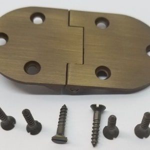 May include: A brass oval hinge with four screw holes on each side. The hinge is closed and has a brushed finish. The image also shows six screws.