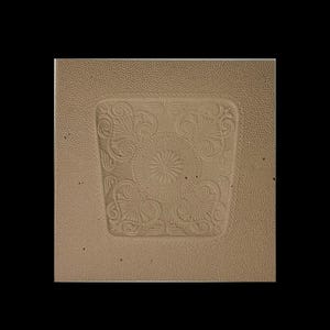 May include: A square, tan-colored object with a textured surface and a central, recessed design. The design features a floral pattern with swirling details and a central sunburst motif. The object is set against a black background.