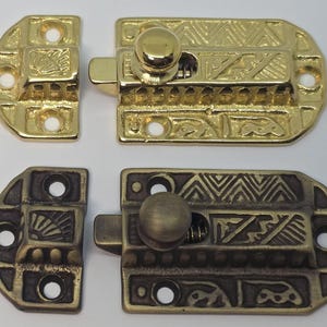 May include: Two decorative cabinet latches, one gold-toned and one bronze-toned, with intricate engraved patterns. Each latch includes a rectangular base and a curved, semi-circular piece. The gold latch is bright and shiny, while the bronze latch has a matte finish.
