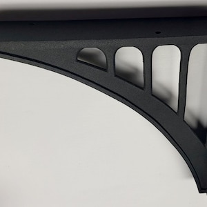 May include: A black, decorative shelf bracket with a curved design. The bracket has a flat top and a series of arched cutouts along its curved edge. It is made of a textured material and is designed to support a shelf.