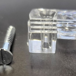 May include: A clear plastic corner bracket with a screw. The bracket has a square shape with a rounded corner. The screw is silver and has a slotted head.