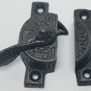 May include: Black cast iron window latch with a decorative design. The latch has a lever handle and a strike plate.