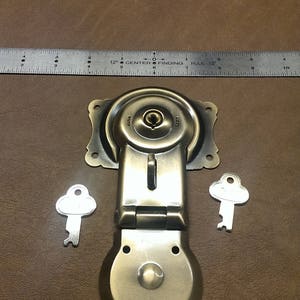 May include: Antique brass latch with two keys. The latch has a round, hinged design with a keyhole and a small knob. The keys are silver and have a simple, rounded design.