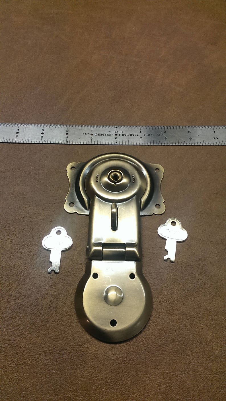May include: Antique brass latch with two keys. The latch has a round, hinged design with a keyhole and a small knob. The keys are silver and have a simple, rounded design.
