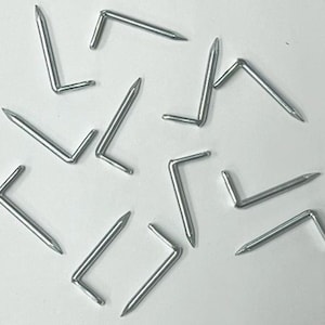 May include: A group of silver-colored L-shaped tacks, also known as upholstery tacks, are scattered on a white surface. The tacks have a sharp point on one end and a bent L-shaped head on the other end.