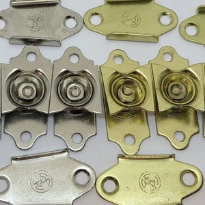 May include: Assortment of metal picture hanging hardware in gold and silver finishes. The hardware features a variety of shapes and sizes, including rectangular, oval, and square.