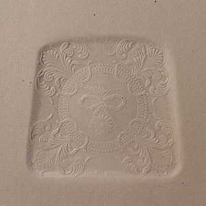 May include: A square, light beige ceramic plate with an embossed design. The design features a central circular emblem with a bow and floral elements, surrounded by ornate scrollwork. The plate's edges are slightly rounded.