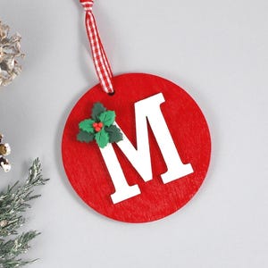 May include: A red wooden Christmas ornament with the letter "M" in white. The ornament has a red and white checkered ribbon for hanging and is decorated with a holly sprig. A festive holiday decoration.