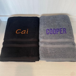 May include: Two folded bath towels, one black and one grey, each featuring a decorative horizontal ribbed band. The black towel is embroidered with the name Cai in orange thread, while the grey towel is embroidered with the name COOPER in purple.