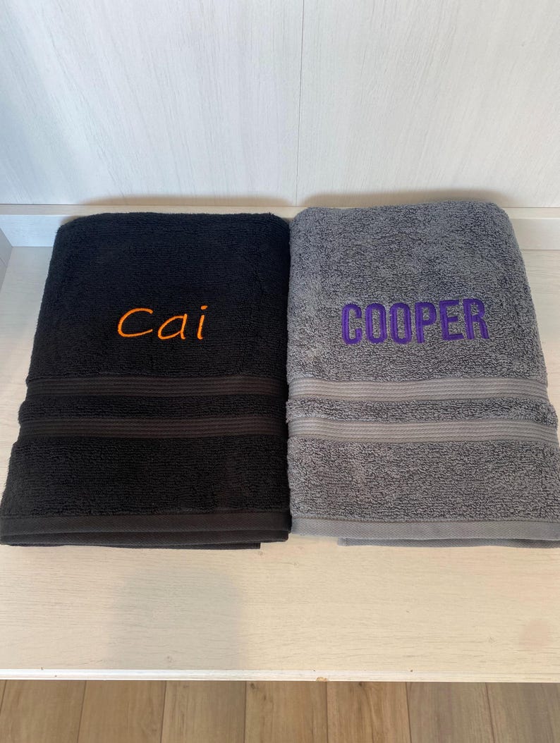 May include: Two folded bath towels, one black and one grey, each featuring a decorative horizontal ribbed band. The black towel is embroidered with the name Cai in orange thread, while the grey towel is embroidered with the name COOPER in purple.