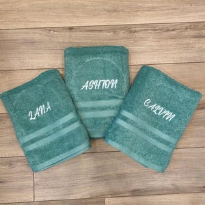 May include: Three teal bath towels featuring embroidered names in white script. The left towel is embroidered with the name Lana, the centre towel with the name Ashton, and the right towel with the name Calvin. Each towel has two horizontal stripes.