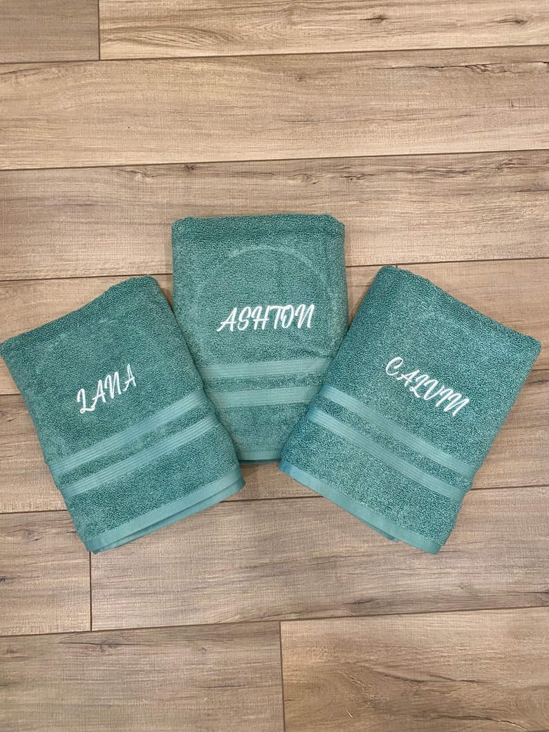 May include: Three teal bath towels featuring embroidered names in white script. The left towel is embroidered with the name Lana, the centre towel with the name Ashton, and the right towel with the name Calvin. Each towel has two horizontal stripes.