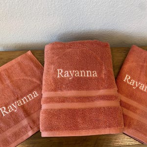 May include: Three coral-colored bath towels with the name "Rayanna" embroidered in white. The towels are folded and arranged on a wooden surface. The towels are a matching set, with a washcloth, hand towel, and bath towel.