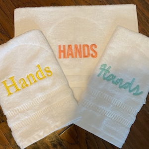 May include: Three white terry towelling cloths featuring embroidered text. One towel displays the word Hands in yellow, another in orange, and the third in light green. Each towel has a textured border with three horizontal lines near the bottom edge.