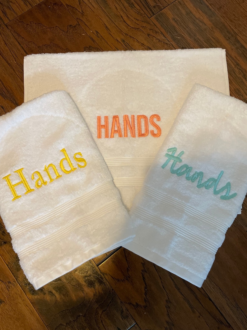 May include: Three white terry towelling cloths featuring embroidered text. One towel displays the word Hands in yellow, another in orange, and the third in light green. Each towel has a textured border with three horizontal lines near the bottom edge.