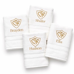 May include: Four white personalized hand towels with gold embroidered crests and names: Brayden, Shafer, Hudson, and Ella. Each towel has a decorative stripe near the bottom.