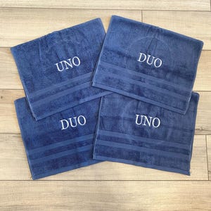 May include: Four rectangular blue towels featuring embroidered white text. Two towels are embroidered with the word UNO and two towels are embroidered with the word DUO. Each towel has a textured surface with three horizontal stripes near the bottom.