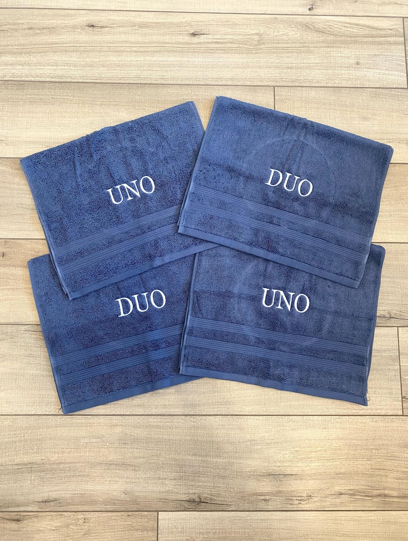 May include: Four rectangular blue towels featuring embroidered white text. Two towels are embroidered with the word UNO and two towels are embroidered with the word DUO. Each towel has a textured surface with three horizontal stripes near the bottom.