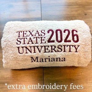 May include: A beige towel featuring maroon embroidery that reads TEXAS STATE UNIVERSITY 2026 and the name Mariana. The text is centred on the folded fabric, displaying a clear, bold font style for the university name and the graduation year.
