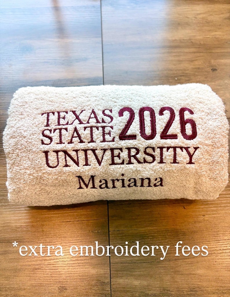 May include: A beige towel featuring maroon embroidery that reads TEXAS STATE UNIVERSITY 2026 and the name Mariana. The text is centred on the folded fabric, displaying a clear, bold font style for the university name and the graduation year.