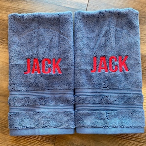 May include: Two rectangular blue terry cloth towels featuring the name JACK embroidered in bold red capital letters across the centre. Each towel has a textured surface with horizontal ribbed bands near the bottom edge and finished hems.